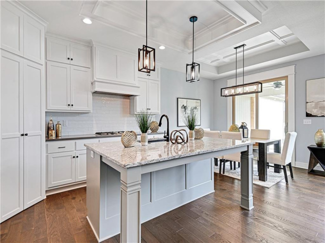 Kitchen with tasteful backsplash, light stone counters, decorative light fixtures, a center island with sink, and white cabinets